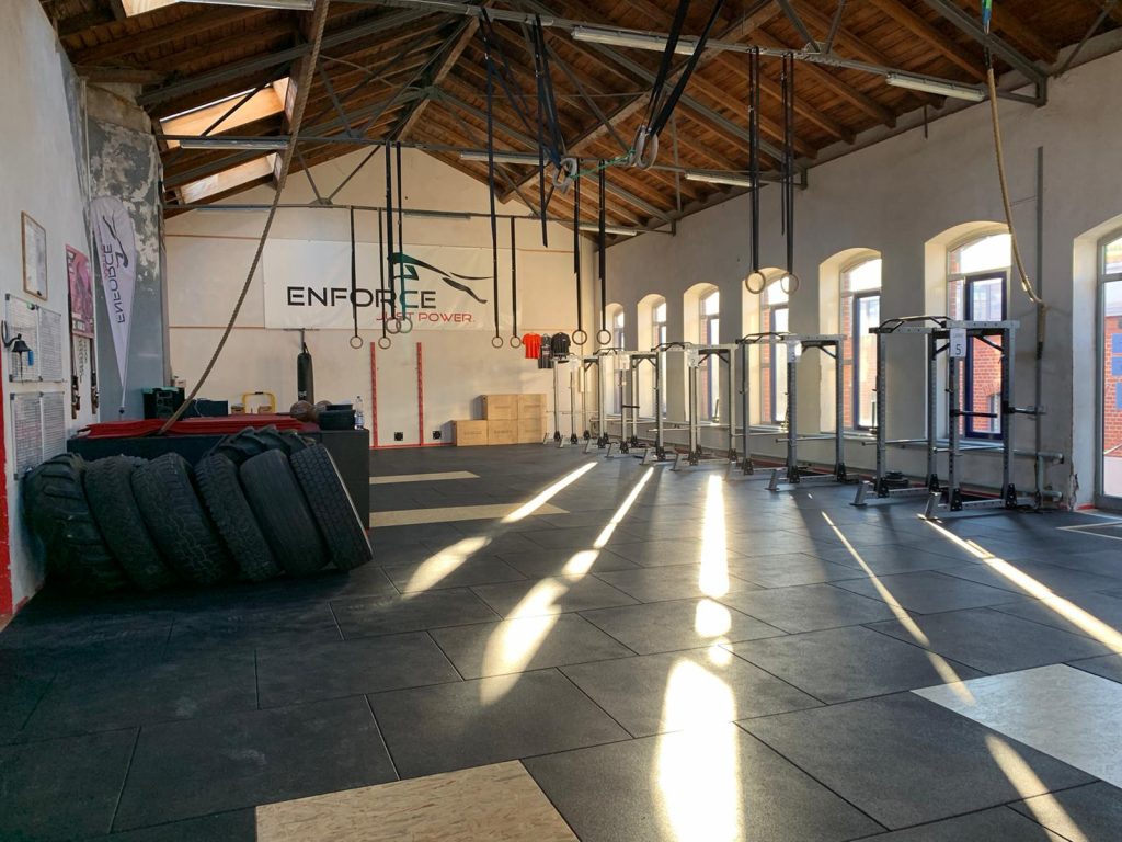 Hier siehst Du die Trainingshalle von ENFORCE-Leipzig. Der Boden ist mit 1 m breiten und rechteckigen Gummifliesen bedeckt. Du siehst zwei darin eingelassene Gewichtheberplattformen aus Holz. AN der rechten Wand sind einige Rundbogenfenster zu sehen, die von 5 Gerätetürmen leicht verdeckt werden. Durch die Fenster strömt warmes Sonnenlicht. Auf der linken Seite stehen mehrere Traktorreifen. Gymnastikringe aus dem Turnen hängen an langen Bändern von der Decke. Die Decke ist aus Holz und ein Spitzdach, ähnlich einem Kirchenschiff gebaut.
